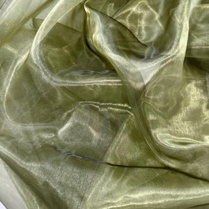Op de afbeelding: Een transparante, lichtgewicht stof in een zachte olijfgroene tint. Het materiaal heeft een uitgesproken doorschijnende kwaliteit met een subtiele, glinsterende glans. Het is gedrapeerd in losse, vloeiende plooien met licht en schaduw.