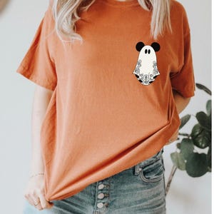 May include: A burnt orange short-sleeve t-shirt featuring a small graphic on the left chest area. The graphic depicts a white ghost character with black mouse-like ears and intricate black floral line art patterns on its body and hem.