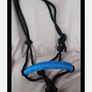 Puede incluir: Un cabezada de cuerda negra con una muserola acolchada azul. La cabezada está hecha de cuerda trenzada y tiene un diseño simple.