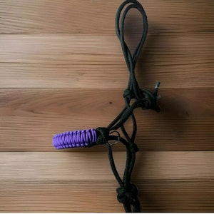 Könnte beinhalten: Ein lila-schwarzes Seilhalfter für Pferde. Das Halfter besteht aus geflochtenem Seil und hat einen Metallring am Nasenband.