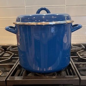 Może przedstawiać: Niebieski emaliowany garnek z pokrywką, stojący na kuchence gazowej. Garnek ma srebrną obręcz i uchwyty. Kuchenka ma czarne ruszty i czerwone pokrętła. Tło z białych płytek.