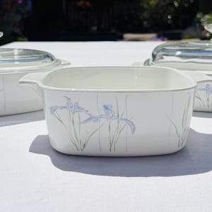 Könnte beinhalten: Drei weiße Keramik-Auflaufformen mit integrierten Griffen und einem Blumenmuster aus hellblauen Irisblüten und grünen Stängeln. Zwei der Formen haben durchsichtige Glasdeckel mit Knauf, eine Form ist offen und ohne Deckel.