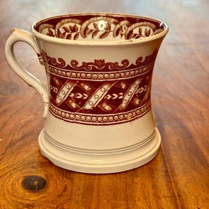 May include: An antique white and maroon ceramic mug with a handle. The mug features a decorative pattern of leaves, swirls, and geometric shapes. The mug has a wide base and a slightly flared top.