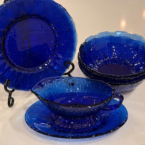 Può includere: Un set di stoviglie in vetro blu cobalto. Il set comprende un piatto grande su un supporto nero, diverse ciotole e una salsiera con un piattino. Il vetro ha un design strutturato e in rilievo.