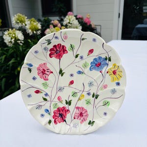 Peut inclure: Une assiette ronde en céramique couleur crème avec un bord festonné, ornée d'un motif floral peint à la main. Le design présente des fleurs rouges, roses, bleues et jaunes reliées par de fines tiges violettes avec des feuilles vertes.