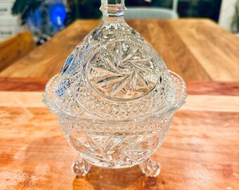 May include: A clear glass candy dish with a matching lid, featuring a pointed finial and intricate pressed patterns including starbursts and diamond cross-hatching. The bowl is supported by three small, curved glass feet at the base.
