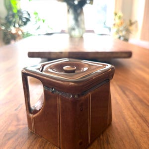 May include: A brown ceramic sugar bowl with a square shape and a handle. The lid has a small knob in the center. Vertical lines decorate the sides of the bowl. The bowl is sitting on a wooden table.
