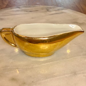 Może przedstawiać: Złoty sosjerka z białym wnętrzem. Sosjerka ma uchwyt z jednej strony i dzióbek z drugiej. Obręcz i zewnętrzna strona są błyszczące w kolorze złotym.