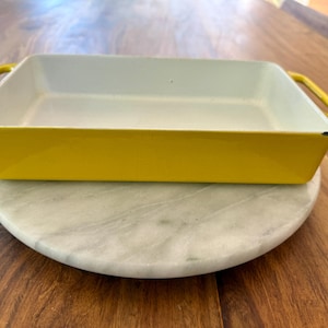 May include: A rectangular yellow baking dish with a white interior and two handles. The dish is sitting on a round, white marble serving board. The dish is designed for baking and serving.