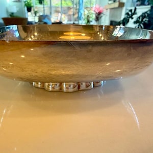 May include: A large, silver-toned decorative bowl with a scalloped base. The bowl has a wide, shallow design with a reflective surface. The rim has a darker, burnished appearance. The bowl is sitting on a light-colored surface.