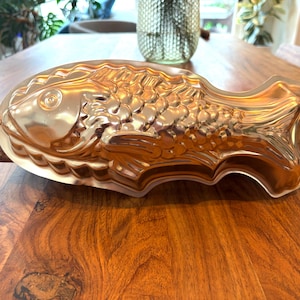 Puede incluir: Un molde vintage con forma de pez, de color dorado. El molde tiene escamas y aletas detalladas, con un asa en un extremo. Está hecho de metal y se apoya sobre una base de madera. Este artículo de cocina está diseñado para hornear o moldear alimentos.