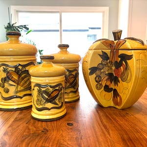 May include: A set of four yellow ceramic kitchen canisters with lids. The canisters feature hand-painted vegetable and fruit designs in brown and black. The largest canister is apple-shaped.