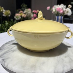 Puede incluir: Un cuenco de cerámica amarillo pálido con tapa a juego. El cuenco tiene dos asas pequeñas y un remolino decorativo en la tapa. El cuenco está sobre una bandeja redonda de mármol.
