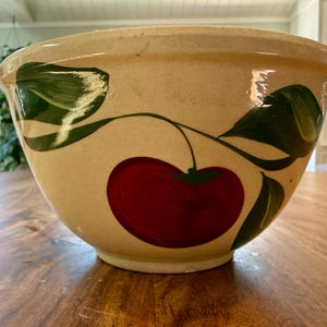 Può includere: Una ciotola in ceramica beige con un grande disegno di ciliegia rossa e foglie verdi. La ciotola ha un bordo largo e una forma arrotondata, adatta per l'uso in cucina.