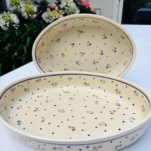 May include: Two oval ceramic baking dishes with a cream-colored base, featuring a repeating pattern of small blue dots and tiny floral clusters in blue and yellow. Each dish has a smooth, glazed finish and a simple, rounded rim design.