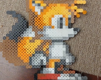 May include: A pixel art bead sprite of the video game character Tails, from the Sonic the Hedgehog franchise. The character is depicted in a standing pose with his two tails extended behind him. The bead sprite is made with a variety of colors, including brown, orange, yellow, white, and black.