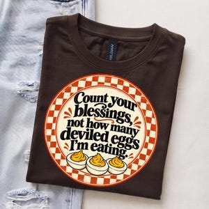 May include: A dark brown t-shirt featuring a circular graphic with a red and white checkered border. The text reads, "Count your blessings, not how many deviled eggs I'm eating," accompanied by an illustration of three deviled eggs.