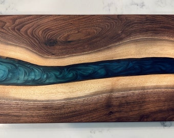 May include: Rectangular wooden serving board with a dark walnut border and a teal epoxy resin river design. The wood grain is visible, and the board is likely used for food preparation or serving.