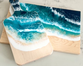 May include: Two wooden cutting boards with a beach-themed epoxy resin design. The resin mimics ocean waves in shades of teal and white, with a sandy beach effect at the base. One board has a handle.