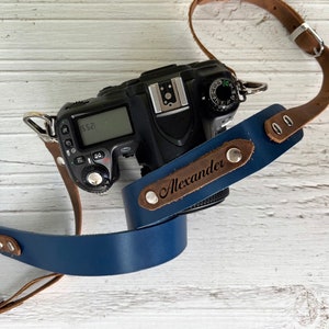 May include: A blue and brown camera strap with the name "Alexander" engraved on it.