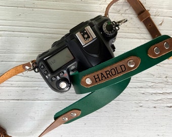 May include: A green leather camera strap with a brown leather trim and the name "HAROLD" embossed in gold lettering. The strap is attached to a black DSLR camera.
