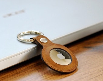 May include: A brown leather keychain with a silver metal ring and a circular metal disc featuring the Apple logo. The keychain is lying on a wooden surface next to a silver laptop.