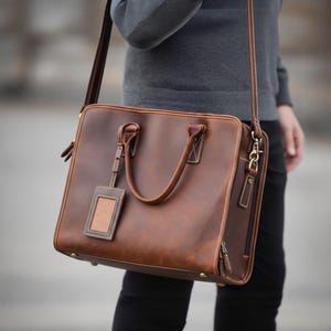 K&ouml;nnte beinhalten: Eine braune Lederaktentasche mit Schulterriemen und oberem Griff. Die Tasche hat eine rechteckige Form mit einem kleinen Anh&auml;nger. Wei&szlig;e N&auml;hte akzentuieren die Kanten und die Tasche wird von einer Person getragen, die einen grauen Pullover und eine schwarze Hose tr&auml;gt.