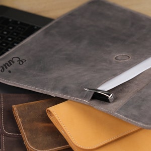 May include: Gray leather document holder with the name "Eunie" engraved on the front. The holder is open, revealing white paper inside. Other leather holders in brown and tan are visible below.