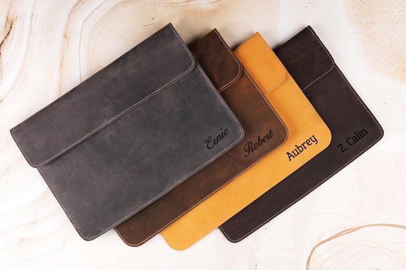 Pu&ograve; includere: Una collezione di custodie per laptop personalizzate in pelle in vari colori: antracite, marrone, beige e marrone scuro. Ogni custodia ha una chiusura a patta ed &egrave; impressa con un nome. Le custodie sono disposte diagonalmente, mostrando i diversi colori e le texture.