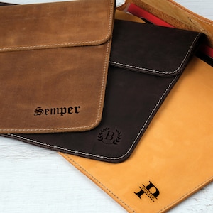 Pu&ograve; includere: Custodie per laptop in pelle nei toni del marrone e del beige, con cuciture bianche. Una custodia &egrave; impressa con la parola "Semper", un'altra con una "B" in una corona e una terza con la lettera "P" e il nome "Porter".