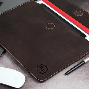 Pu&ograve; includere: Una cartella in pelle marrone scuro con cuciture bianche, aperta per rivelare documenti e un portadocumenti rosso. Un laptop, un mouse bianco e una penna sono accanto alla cartella. La cartella ha le iniziali "KL" in un cerchio.