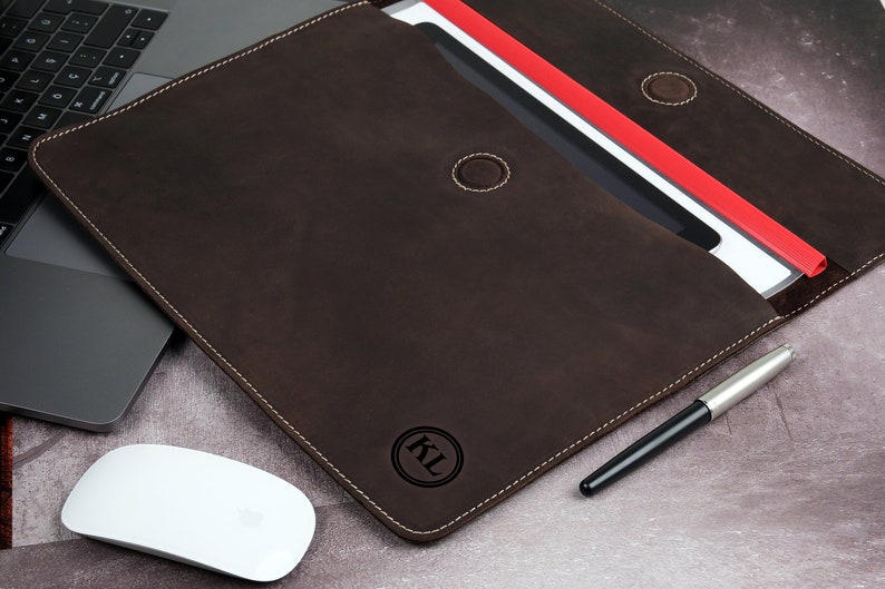 Pu&ograve; includere: Una cartella in pelle marrone scuro con cuciture bianche, aperta per rivelare documenti e un portadocumenti rosso. Un laptop, un mouse bianco e una penna sono accanto alla cartella. La cartella ha le iniziali "KL" in un cerchio.