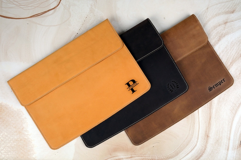 Pu&ograve; includere: Tre custodie per laptop in pelle color cuoio, nero e marrone. Ogni custodia ha una chiusura a patta ed &egrave; personalizzata con un monogramma o un nome. La custodia color cuoio ha la lettera "P" e il nome "Porter". La custodia nera ha una corona d'alloro e la custodia marrone ha il nome "Semper".
