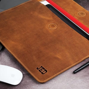 Pu&ograve; includere: Una custodia per laptop in pelle marrone con le iniziali "JED" in rilievo sul davanti. La custodia &egrave; aperta, rivelando un tablet e un documento rosso. Un mouse bianco e una penna sono accanto alla custodia.