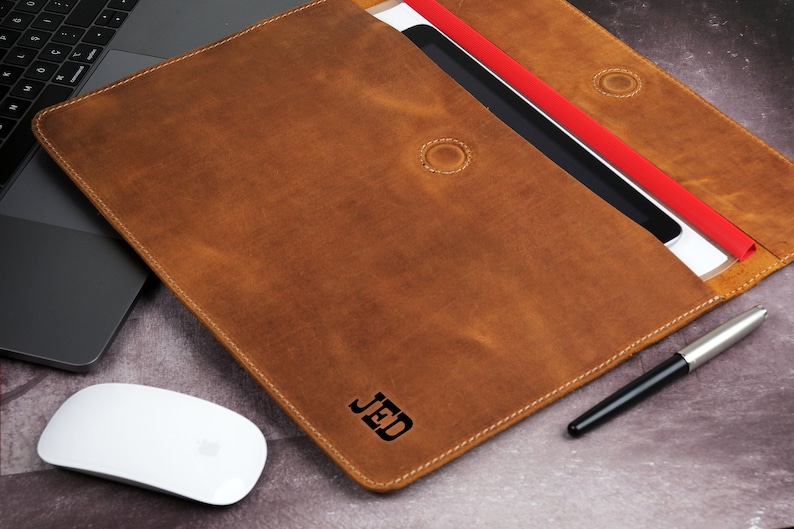 Pu&ograve; includere: Una custodia per laptop in pelle marrone con le iniziali "JED" in rilievo sul davanti. La custodia &egrave; aperta, rivelando un tablet e un documento rosso. Un mouse bianco e una penna sono accanto alla custodia.