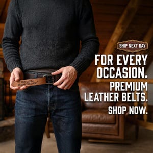 Può includere: Una cintura in pelle marrone scuro con fibbia in metallo color argento, caratterizzata dalla scritta "YOUR CUSTOM MESSAGE GOES HERE" incisa sul lato interno del cinturino. La cintura è indossata con jeans blu scuro e un maglione nero a maniche lunghe.