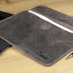 Pu&ograve; includere: Una custodia per laptop in pelle grigia con il nome "Ernie" inciso sul davanti. La custodia &egrave; aperta, rivelando un interno bianco e un accento marrone. Una penna nera &egrave; appoggiata accanto alla custodia.
