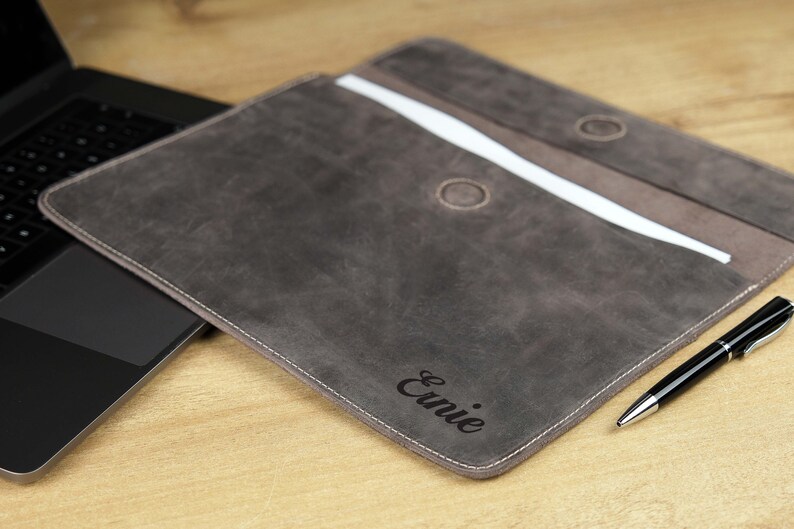 Pu&ograve; includere: Una custodia per laptop in pelle grigia con il nome "Ernie" inciso sul davanti. La custodia &egrave; aperta, rivelando un interno bianco e un accento marrone. Una penna nera &egrave; appoggiata accanto alla custodia.