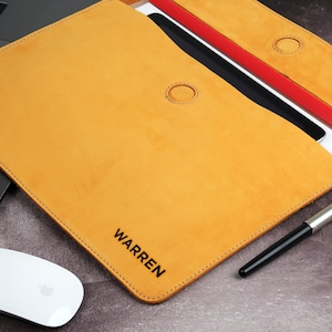 May include: Mustard-colored leather laptop sleeve with the name "WARREN" printed in black. The sleeve is open, revealing a tablet and papers. A silver and black pen and a white mouse are also visible.