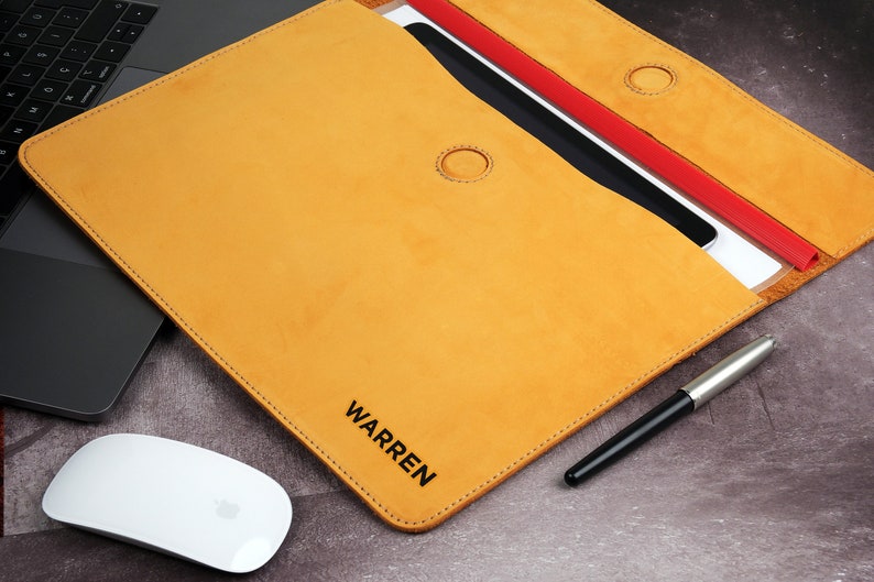 Pu&ograve; includere: Custodia per laptop in pelle color senape con il nome "WARREN" stampato in nero. La custodia &egrave; aperta, rivelando un tablet e dei fogli. Sono visibili anche una penna argento e nera e un mouse bianco.