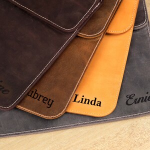 Pu&ograve; includere: Primo piano di quattro custodie in pelle per tablet nei toni del marrone, beige e grigio. Ogni custodia &egrave; personalizzata con un nome in corsivo. Cuciture bianche delineano i bordi.