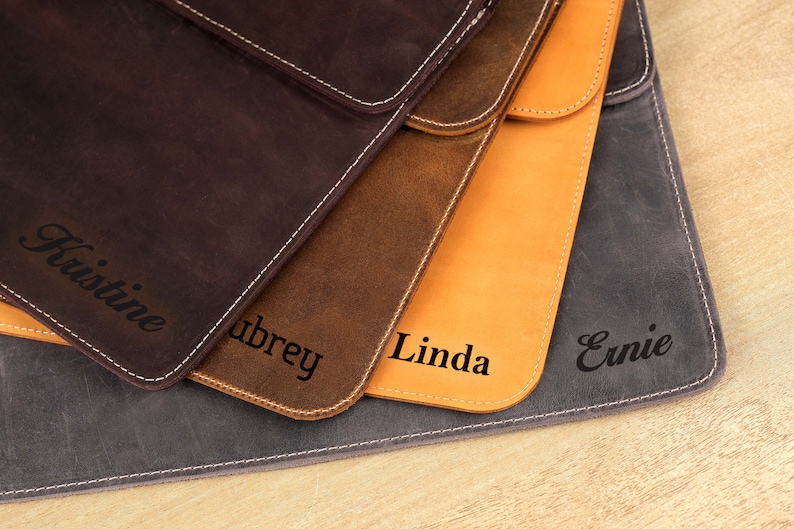 Pu&ograve; includere: Primo piano di quattro custodie in pelle per tablet nei toni del marrone, beige e grigio. Ogni custodia &egrave; personalizzata con un nome in corsivo. Cuciture bianche delineano i bordi.