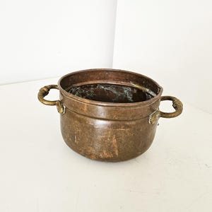 Op de afbeelding: Een vintage koperen pot met twee messing handvatten. De pot heeft een afgeronde body en een licht uitlopende rand. Het koper heeft een rijke, verouderde patina en de messing handvatten vertonen tekenen van slijtage. Dit is een decoratief keukenartikel.
