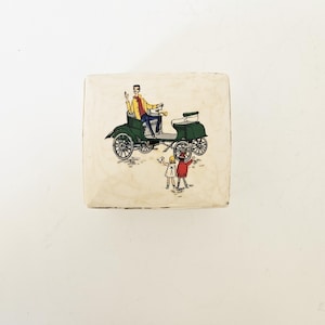 Op de afbeelding: Vierkante, crèmekleurige keramische doos met een vintage illustratie van een groene auto met een bestuurder in een gele jas en twee kinderen. De auto heeft zwarte wielen en de kinderen dragen rode en witte jurken.