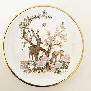 Op de afbeelding: Een decoratieve schotel met een gouden rand en een witte achtergrond. De schotel heeft een gedetailleerde illustratie van twee herten onder een boom met roze bloemen. Het ontwerp bevat een vlinder en een gebarsten, verouderde uitstraling.