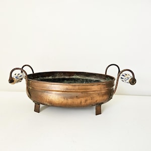 Peut inclure: Un bol en métal cuivré de forme arrondie, soutenu par trois petits pieds. Le bol est doté de deux poignées incurvées avec des extrémités décoratives en céramique à motifs floraux bleus et blancs. L'intérieur du bol montre des signes d'âge.