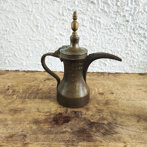 Könnte beinhalten: Eine Dallah-Kaffeekanne aus Messing mit breitem Boden, geschwungenem Griff, langem, spitz zulaufendem Ausguss und einem abnehmbaren Deckel mit dekorativem Knauf. Die Metalloberfläche weist eine verwitterte Patina mit sichtbaren Altersspuren auf.
