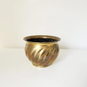 Könnte beinhalten: Ein Vintage-Blumentopf aus Messing mit einem wirbelnden, strukturierten Design. Der Pflanztopf hat eine breite, abgerundete Form mit einem ausgestellten Rand und einem dunklen Innenraum. Das Messing hat eine warme, gealterte Patina, die seinen antiken Charme unterstreicht. Ein dekoratives Stück für Zimmerpflanzen.