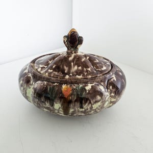 Op de afbeelding: Een keramische pot met deksel met een gespikkeld bruin en wit patroon. De pot heeft een afgeronde vorm met een decoratieve knop op het deksel. De basis heeft groene en oranje bloemaccenten.