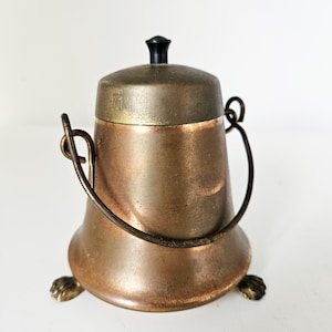 Op de afbeelding: Een vintage metalen suikerpot met een deksel en handvat. De kom heeft een koperkleur met een messingkleurig deksel en een zwarte knop. Het heeft drie kleine klauwvoeten.